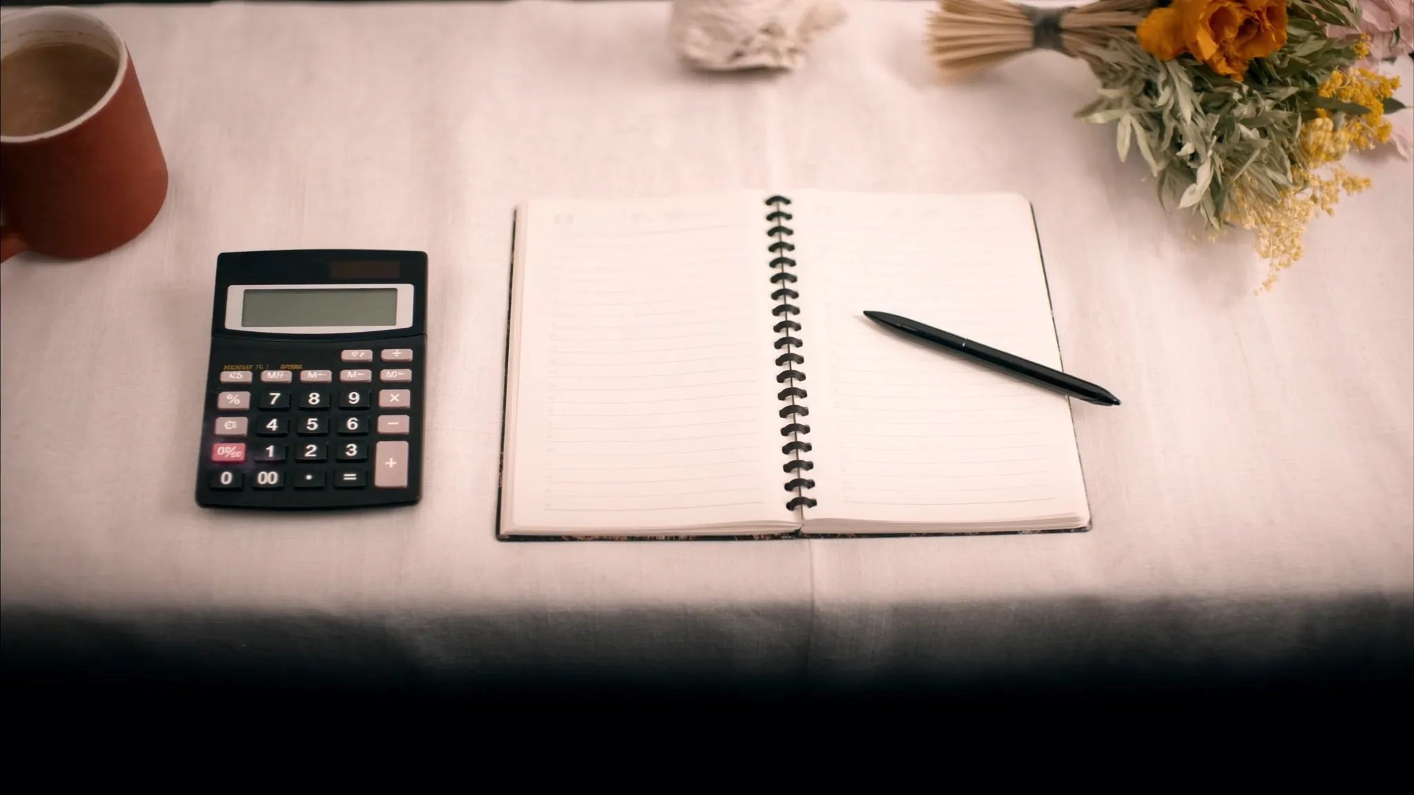 A wedding budget flat-lay with a calculator, notebook, coffee and dried flowers