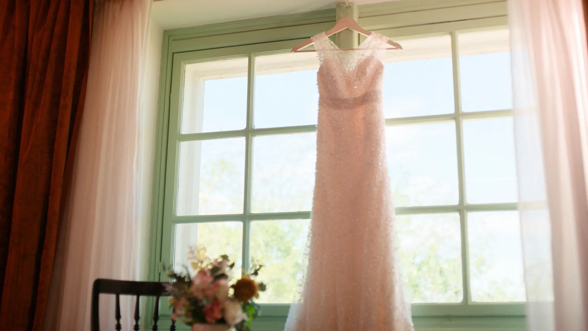 A beaded wedding dress on a hanger by a bright window in soft morning light